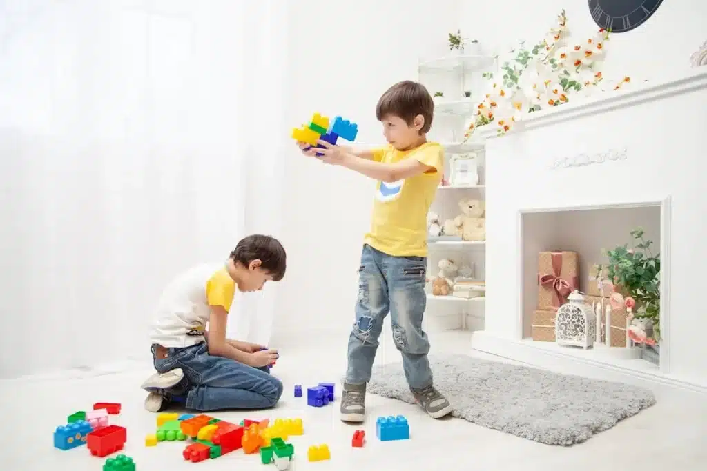 Duas crianças brincam no chão de uma sala de estar com blocos de montar coloridos. Uma criança monta uma estrutura, e a outra observa, representando atividades divertidas para fazer no tédio.