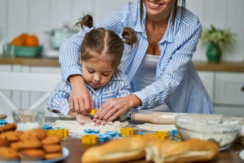 Mãe e filha pequenas, vestindo camisas listradas combinando, trabalham juntas na cozinha para cortar formas de cookies da massa aberta. A menina está concentrada enquanto a mãe guia suas mãos. Cortadores de biscoito coloridos estão espalhados pela mesa, junto com um rolo de massa, farinha e biscoitos já assados em um prato ao lado. A imagem captura um momento doce e educativo sobre "como fazer cookies" com participação infantil.