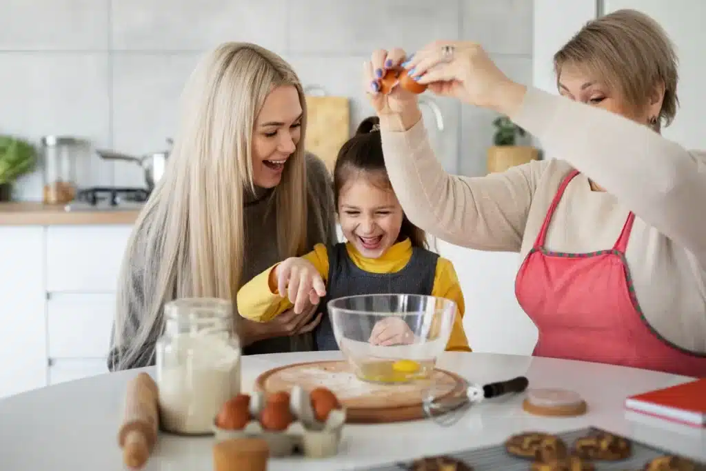 Três gerações de mulheres, avó, mãe e filha, riem e se divertem juntas na cozinha enquanto preparam a massa de cookies. A avó quebra um ovo na tigela, a criança sorri animada, e a mãe observa com carinho. A mesa de cozinha está cheia de ingredientes como farinha, ovos e biscoitos prontos na grade de resfriamento. A imagem ilustra o tema "como fazer cookies" de uma forma alegre e familiar.