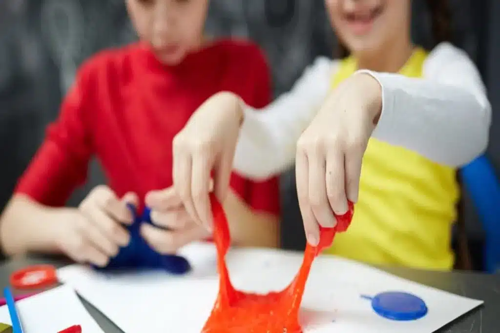 Duas crianças brincando com slime vermelho e azul, esticando a massa em uma mesa de trabalho. A imagem ilustra o processo e a diversão de como fazer slime caseiro.