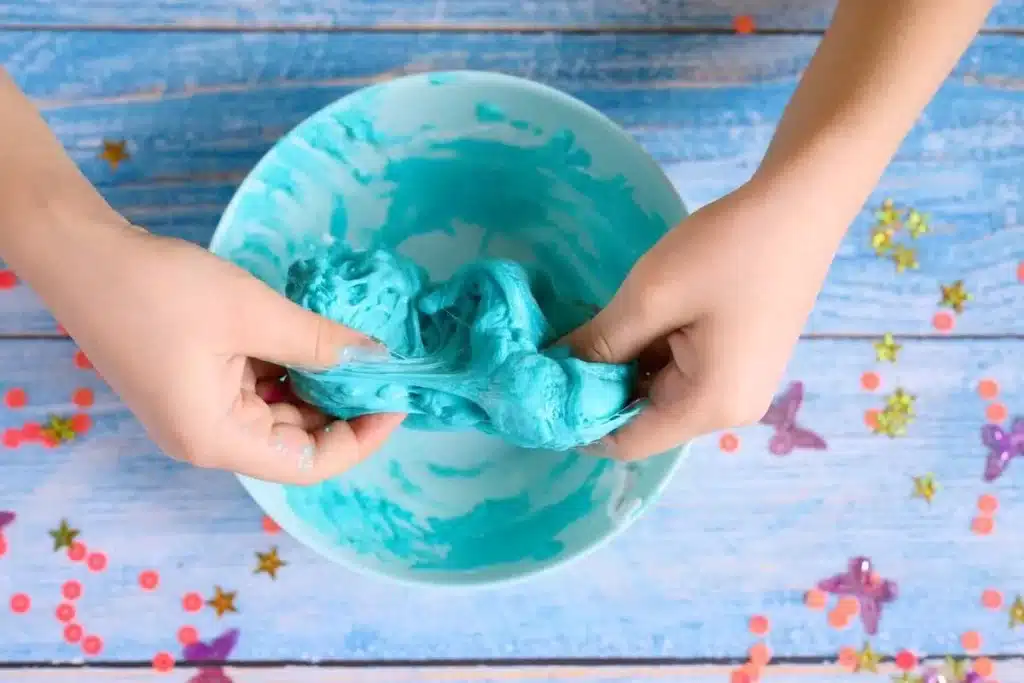 Duas mãos de criança misturam uma massa de slime azul-turquesa em uma tigela, sobre uma mesa de madeira azul decorada com confetes coloridos, ilustrando como fazer slime caseiro.