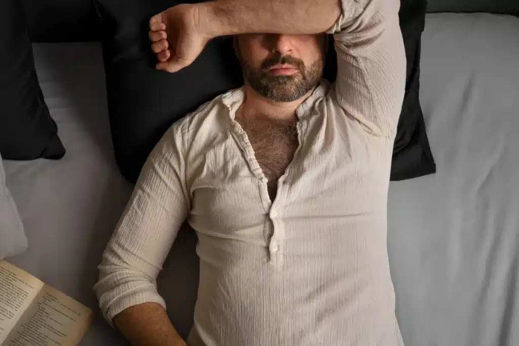 Um homem deitado em uma cama com o braço sobre os olhos, parecendo estressado ou exausto, representando a preocupação de ter uma 'criança perdida' e a angústia parental.