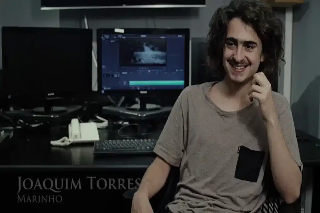  Joaquim Torres Marinho, filho de Fernanda Torres, sentado em uma cadeira em frente a um computador e sorrindo.