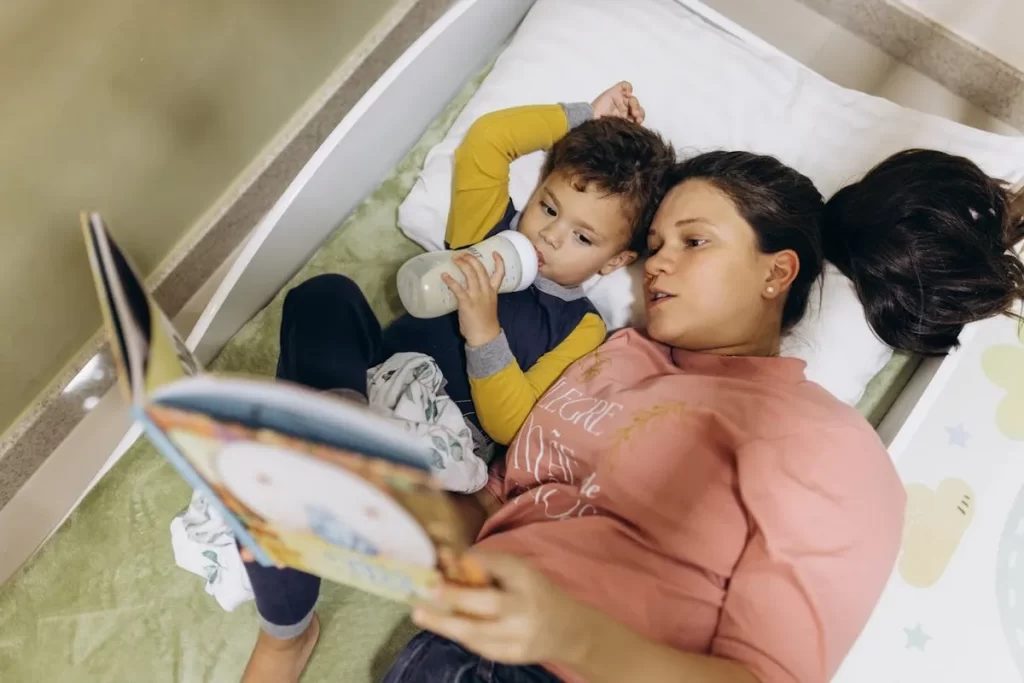Uma mãe deitada com seu filho, que bebe mamadeira, enquanto ela lê um livro de história infantil educativa para ele na cama.