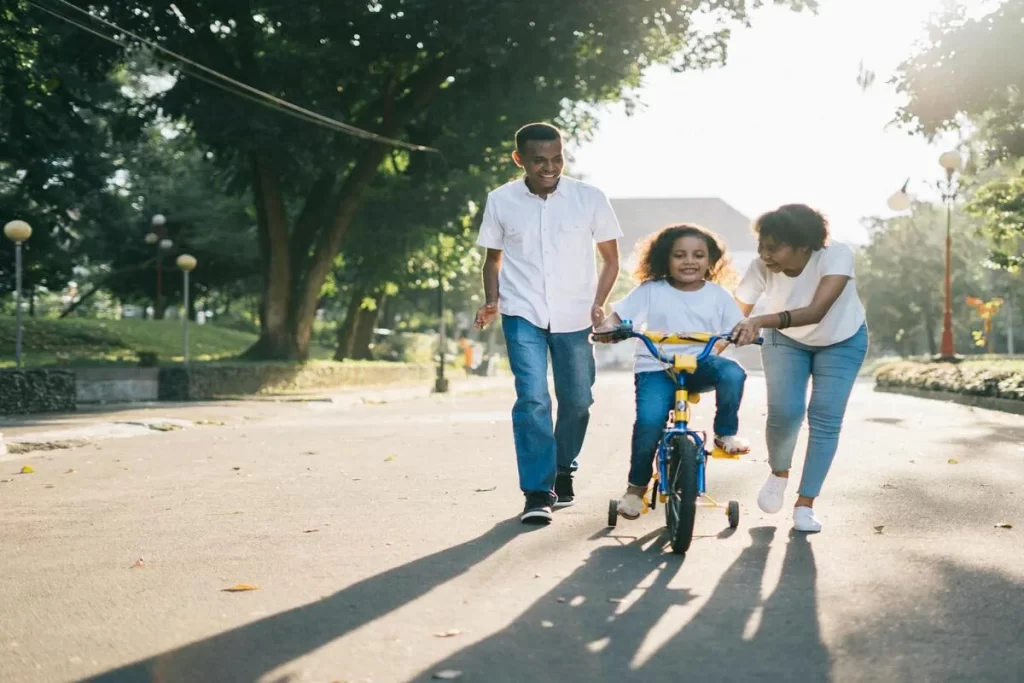 Pai e mãe, sorrindo, ensinam sua filha a andar de bicicleta em um parque em um dia ensolarado.