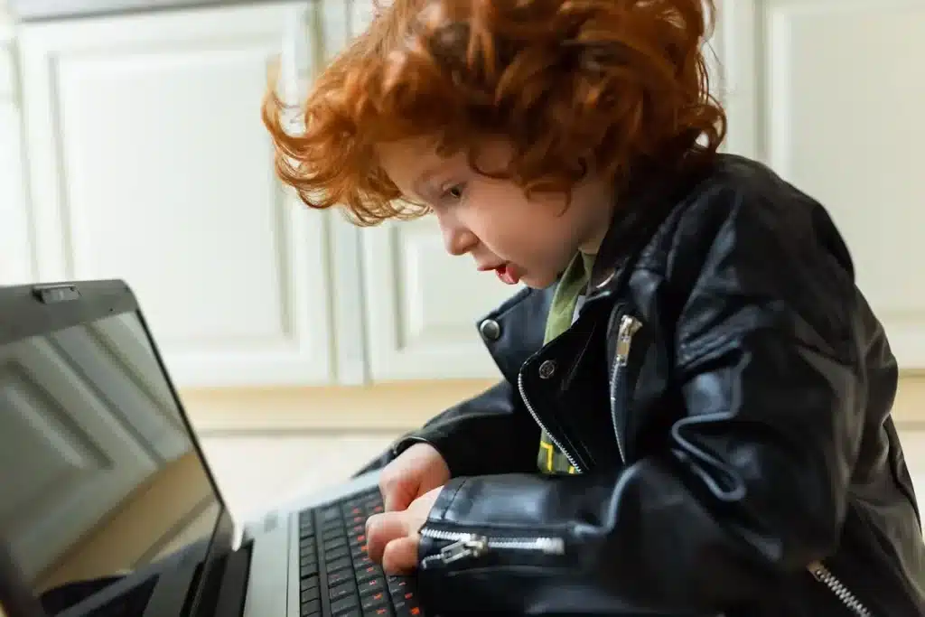 Um menino ruivo, de cabelo cacheado, usando uma jaqueta de couro ecológico preta, olha para um laptop. A imagem destaca a jaqueta infantil para menino em um contexto moderno e casual.