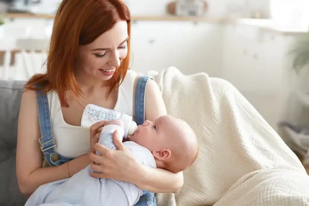Mulher com cabelo ruivo sorrindo e segurando um bebê que se alimenta de uma mamadeira, representando a nutrição infantil.