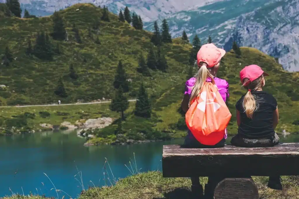 Duas crianças, com bonés e mochilas, sentadas de costas em um banco de madeira, observando um lago e montanhas, representando lugares para viajar com crianças e apreciar a natureza.