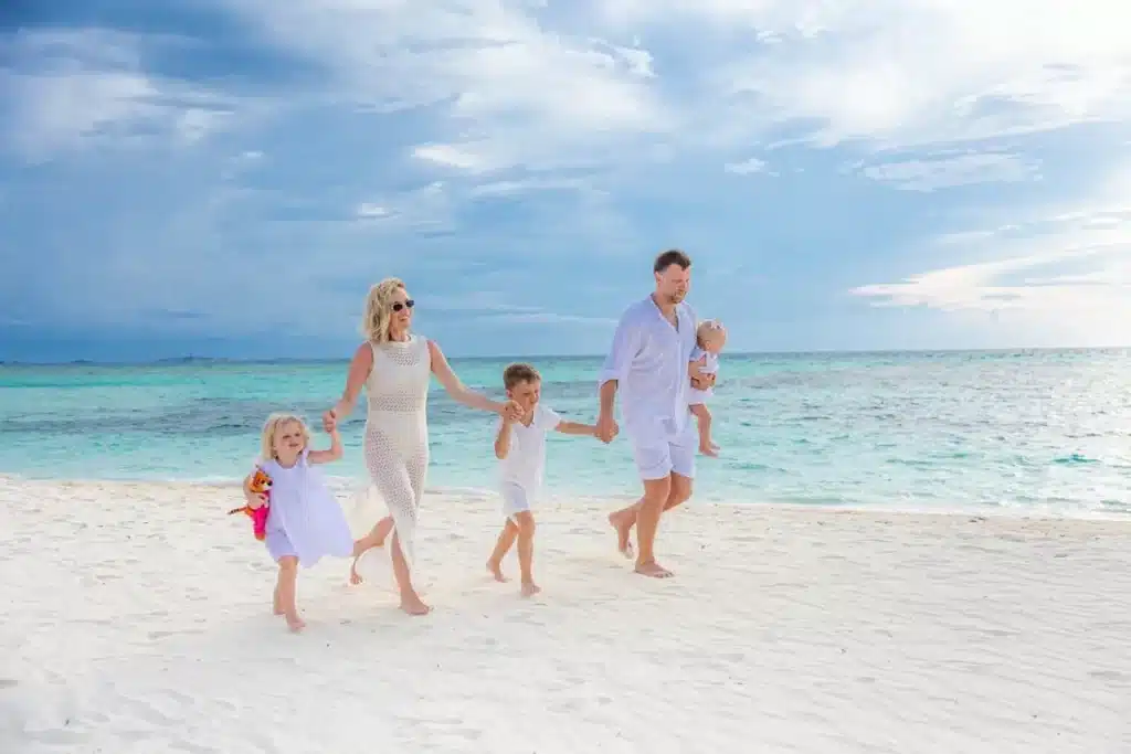 Uma família, com dois pais e três crianças, caminha de mãos dadas em uma praia de areia branca e água azul-turquesa, representando lugares ideais para viajar com crianças.