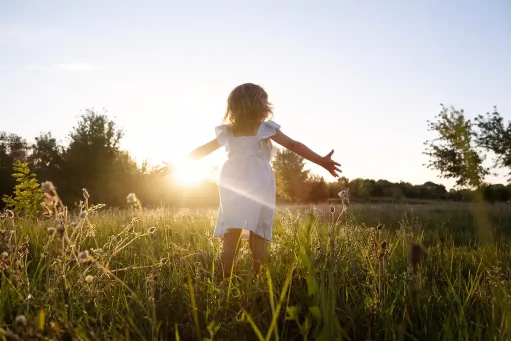 Criança com braços abertos em um campo de grama alta, sentindo a luz do sol poente, simbolizando a liberdade, a alegria e a busca por uma conexão espiritual.