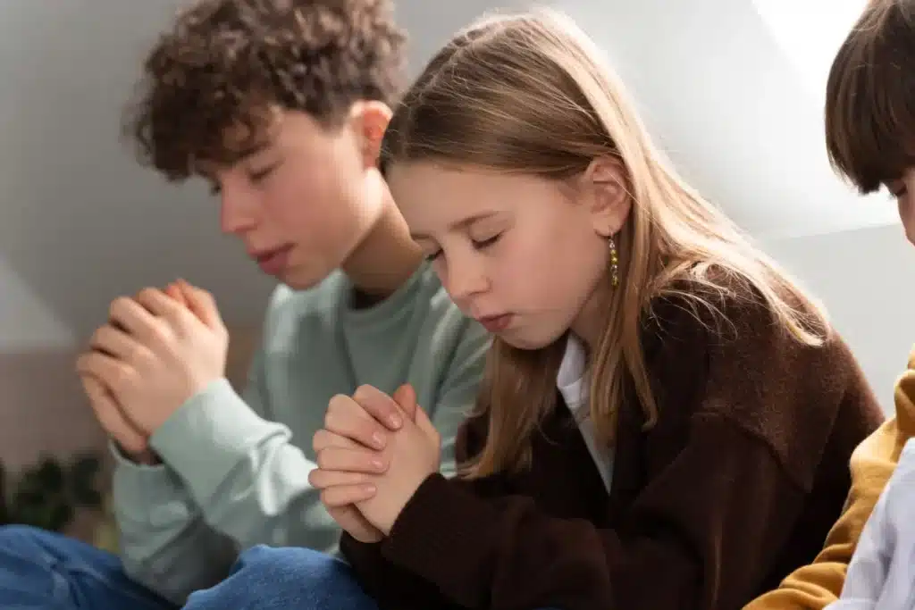 Crianças com as mãos unidas e olhos fechados em oração, refletindo sobre fé e o significado do que é Deus.