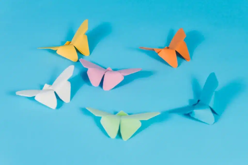 Seis borboletas de origami coloridas em tons de amarelo, laranja, rosa, branco, verde e azul, flutuando sobre um fundo azul claro.