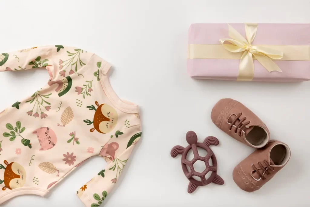 Uma vista superior de itens para recém-nascido, incluindo um macacão rosa com estampa de cervos, sapatos de bebê, um mordedor de tartaruga e uma caixa de presente. A imagem sugere um kit de boas-vindas completo e delicado para o bebê.