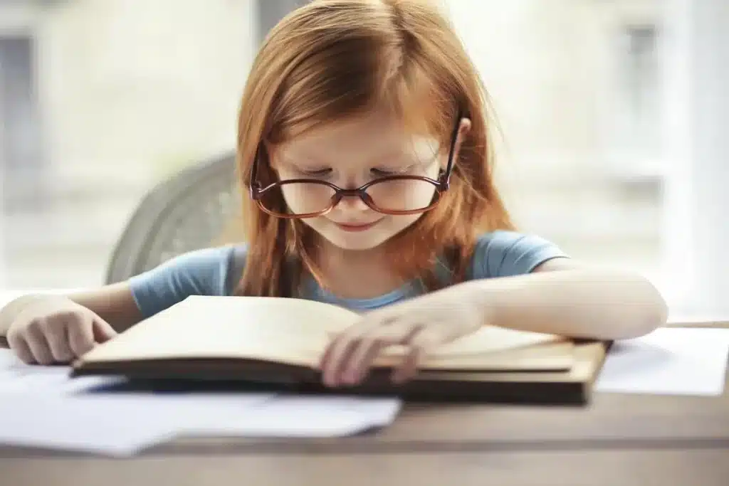 Menina com óculos grandes lendo um livro em uma mesa, representando a saúde ocular.