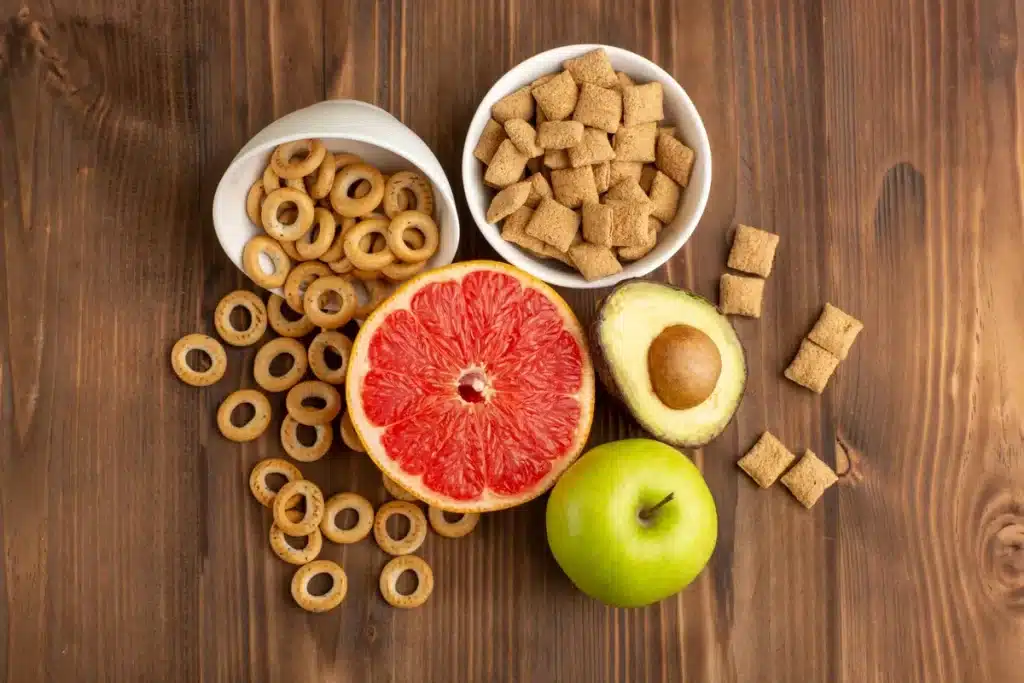 Snacks saudáveis, incluindo frutas como maçã e toranja, além de abacate e biscoitos integrais, dispostos sobre uma mesa de madeira.
