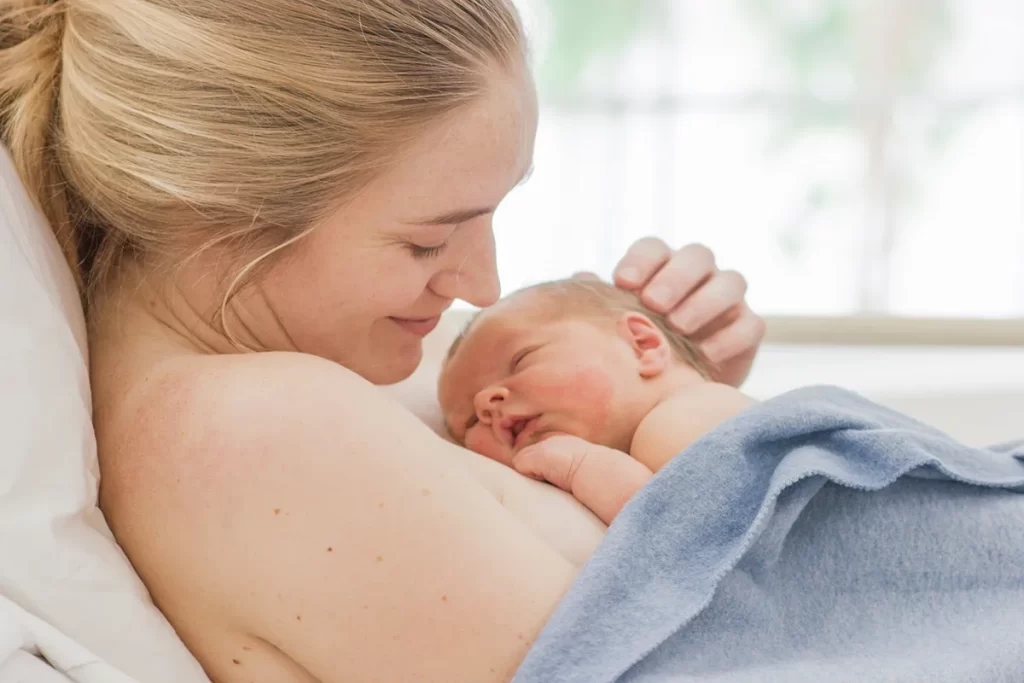 Uma mãe feliz e sorridente, com cabelo loiro preso, está deitada na cama, abraçando seu bebê recém-nascido. O bebê, com os olhos fechados, está aninhado no peito da mãe, que o segura com uma das mãos na cabeça, enquanto ele está parcialmente coberto por um cobertor azul. A imagem transmite intimidade, amor e o forte vínculo criado pelo contato pele a pele.