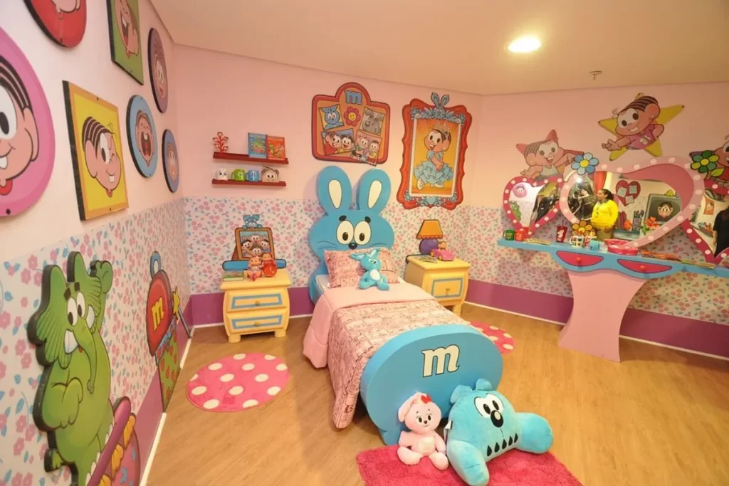 Um quarto temático do Parque da Mônica com paredes cor-de-rosa decoradas com os personagens da Turma da Mônica. A cama principal tem uma cabeceira em forma do personagem Bidu, com brinquedos de pelúcia no chão e sobre a cama.Esta fotografia exibe um quarto temático do Parque da Mônica, projetado para imergir os visitantes no universo dos quadrinhos de Maurício de Sousa. O ambiente é dominado por uma paleta de cores alegre, com paredes em um tom suave de rosa e estampas florais.  A cama é o destaque principal, com uma cabeceira criativa em forma do personagem Bidu. O espaço é enriquecido com recortes de madeira dos principais personagens, como Cebolinha, Mônica e Cascão, que estão fixados nas paredes. No chão e sobre a cama, bonecos de pelúcia dos personagens, como o coelho azul e o cão Bidu, completam a atmosfera lúdica.  A imagem mostra como a marca Turma da Mônica se estende além das atrações do parque, criando uma experiência completa para o público infantil. A penteadeira em forma de coração e a iluminação suave contribuem para o ambiente de sonho.