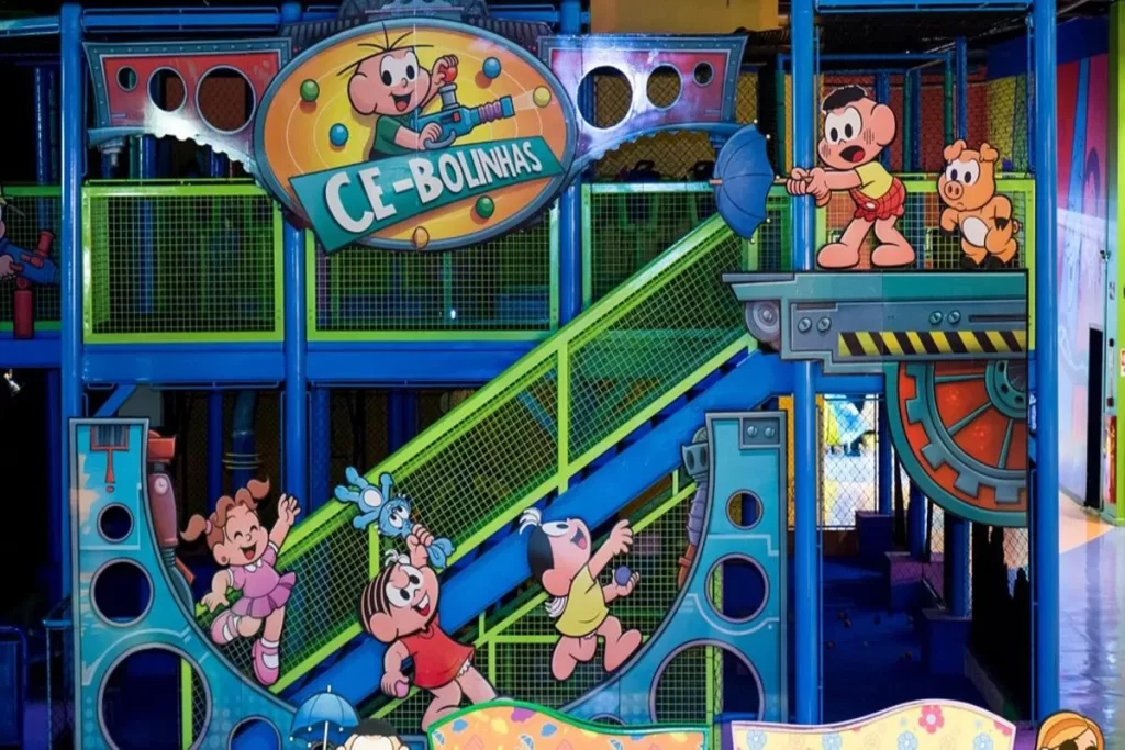 Um grande playground colorido, em área indoor, do Parque da Mônica. A estrutura é decorada com ilustrações dos personagens da Turma da Mônica, como Cebolinha, Mônica, Cascão e Magali, e tem uma placa que diz "CE-BOLINHAS".