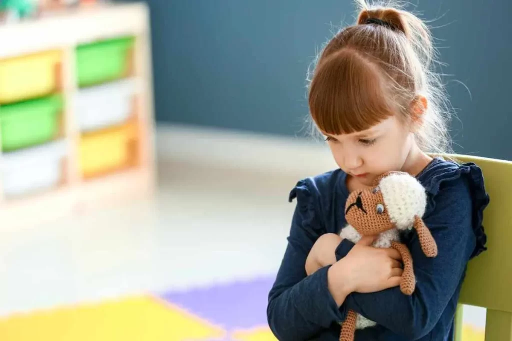 Uma menina com rabo de cavalo, sentada e abraçando um pequeno brinquedo de pelúcia, com uma expressão triste ou introspectiva. A imagem ilustra temas de emoções infantis e terapia (TPS), com um fundo de sala de jogos.