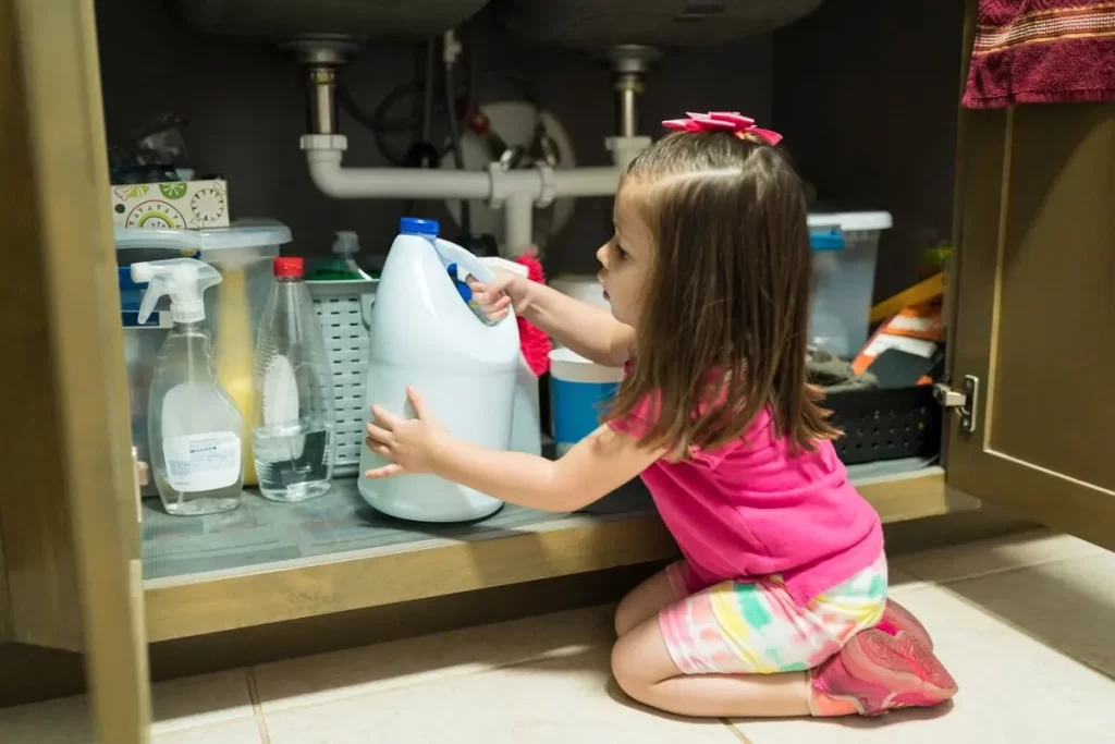 Menina pequena brincando com produtos de limpeza sob a pia da cozinha, representando um acidente doméstico.