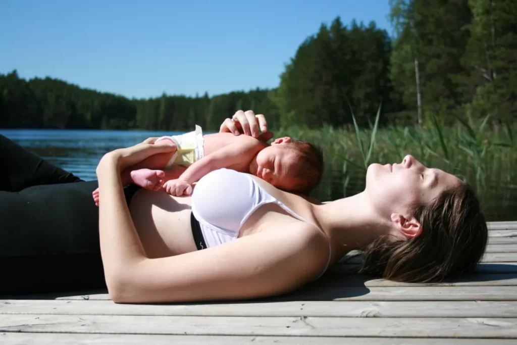 Mãe relaxando com seu bebê recém-nascido deitado em seu peito em um deque de madeira sob o sol.