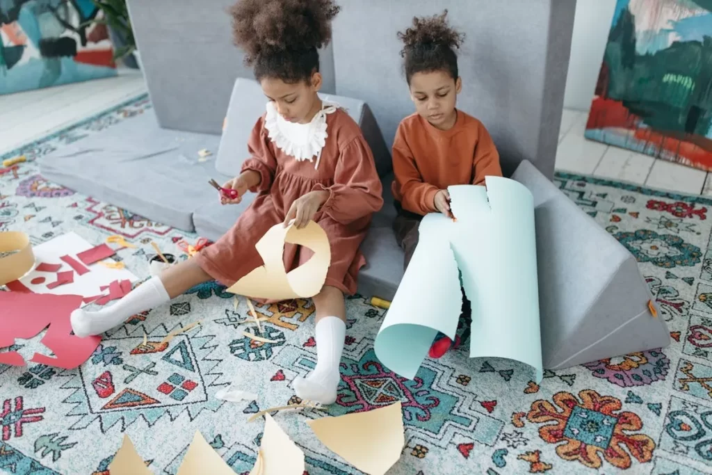 Duas meninas pequenas em um tapete fazendo artesanato com papel, tesouras e colagem.