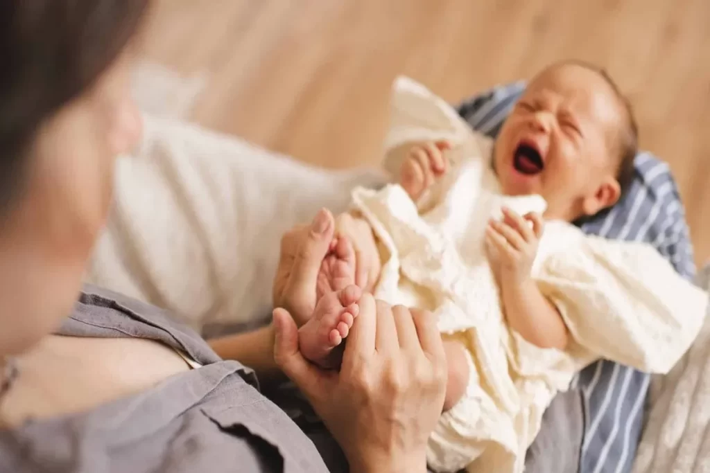 Mãe ou cuidador segurando os pés de um bebê que chora, com o rosto franzido e a boca aberta, expressando dor ou desconforto.