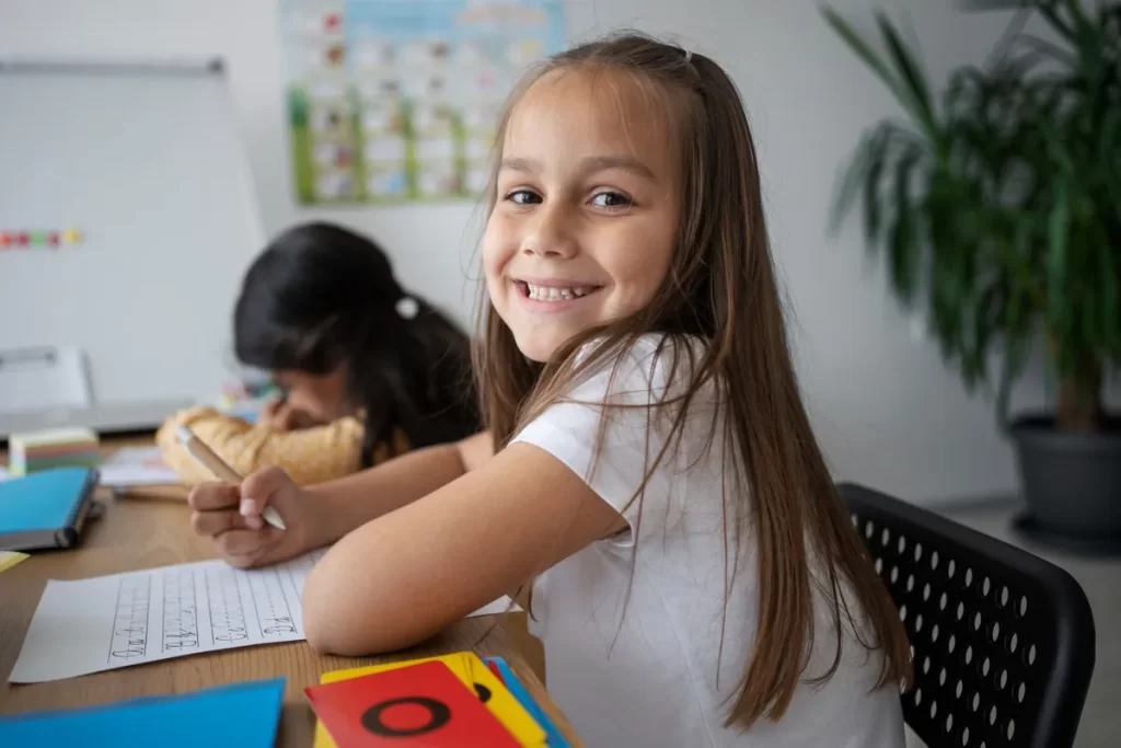 Uma menina sorrindo enquanto estuda inglês em sala de aula, com outra criança ao fundo.
