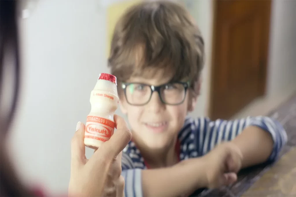 Mão adulta oferecendo um frasco de Yakult para um menino sorridente, simbolizando a saúde da microbiota intestinal.