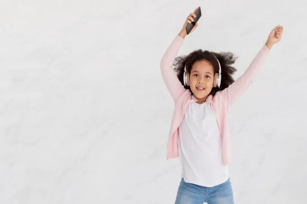 Menina criança, com cabelo cacheado e pele morena, em um suéter rosa e jeans, dançando e pulando com os braços levantados, enquanto ouve músicas infantis em um fone de ouvido branco e segura um smartphone. O fundo é branco e liso.
