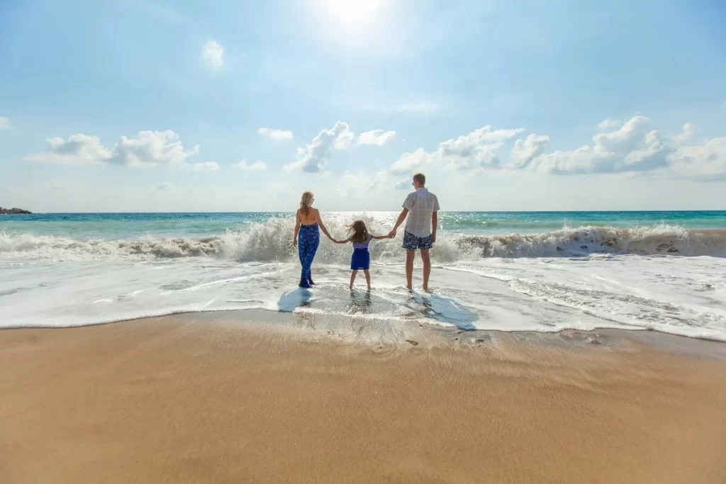 Uma família de três pessoas, de costas, de mãos dadas, à beira da praia, observando o mar em um dia de sol. O pai e a mãe estão de cada lado, com a filha no meio. A água do mar chega perto de seus pés e a areia da praia está em primeiro plano.