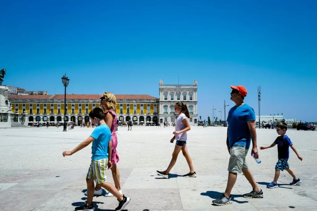 Uma família de cinco pessoas — mãe, pai e três crianças — caminhando em uma praça pública em um dia de sol. A imagem captura a alegria e a união familiar, destacando o pai com um boné laranja e as crianças.