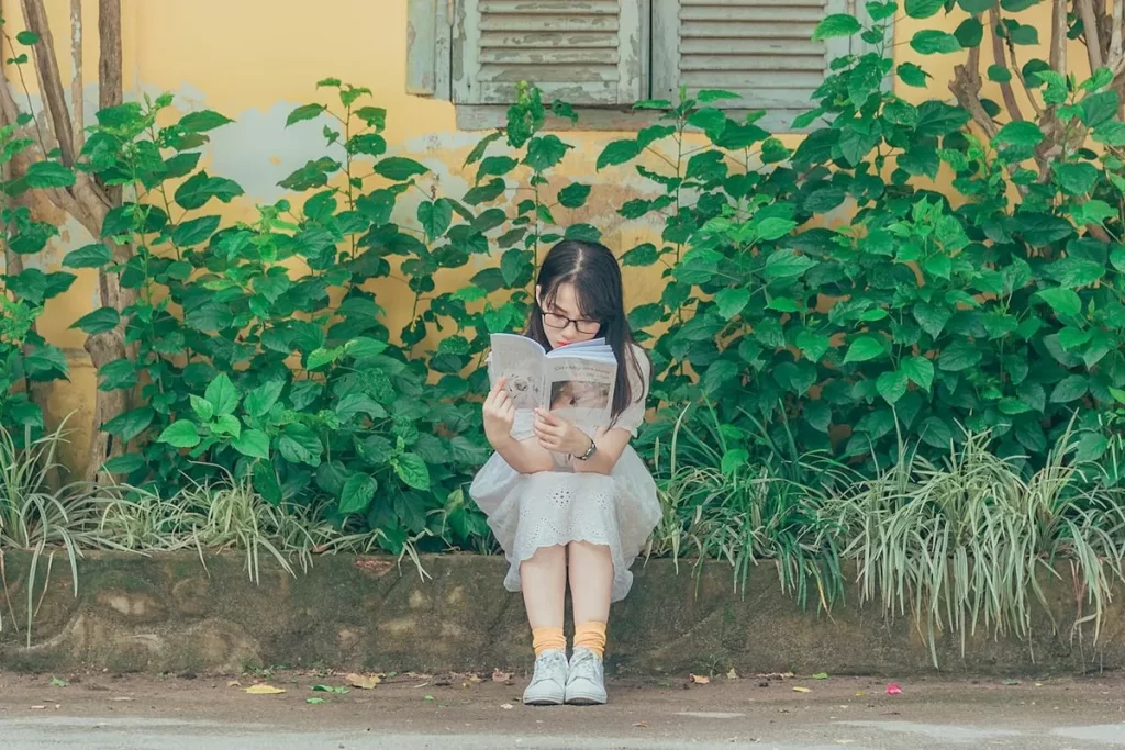 Menina com óculos sentada em um muro lendo um livro em um jardim com folhagens verdes.
