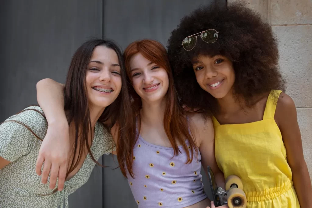 Três amigas pré-adolescentes de etnias diferentes sorriem e posam juntas. Elas vestem roupas casuais de verão, incluindo um vestido estampado, uma blusa com girassóis e uma blusa amarela, enquanto uma delas segura um skate.
