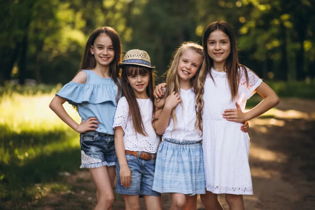 Quatro meninas pré-adolescentes, de diferentes idades e alturas, sorriem e posam juntas ao ar livre. Elas vestem roupas femininas casuais e de verão, incluindo shorts jeans, blusas claras, um vestido de renda branco e um chapéu de palha.