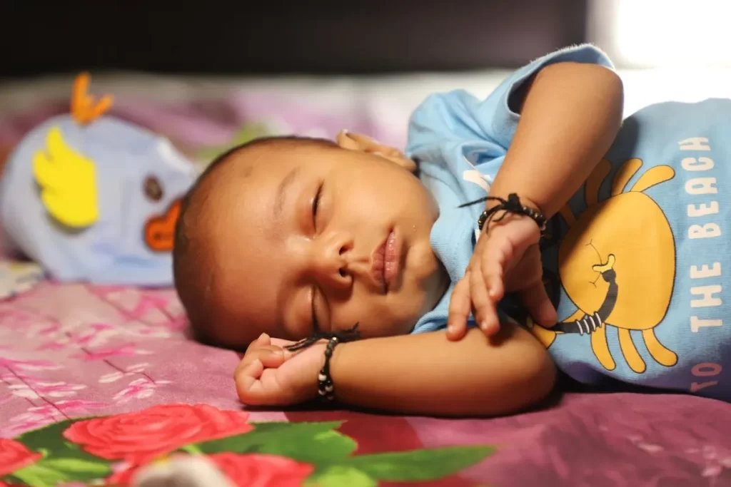 Bebê recém-nascido dormindo de lado em uma cama. Ele usa uma camiseta azul com estampa de caranguejo e, ao seu lado, há um brinquedo macio de pato. A imagem captura a serenidade de um bebê dormindo.