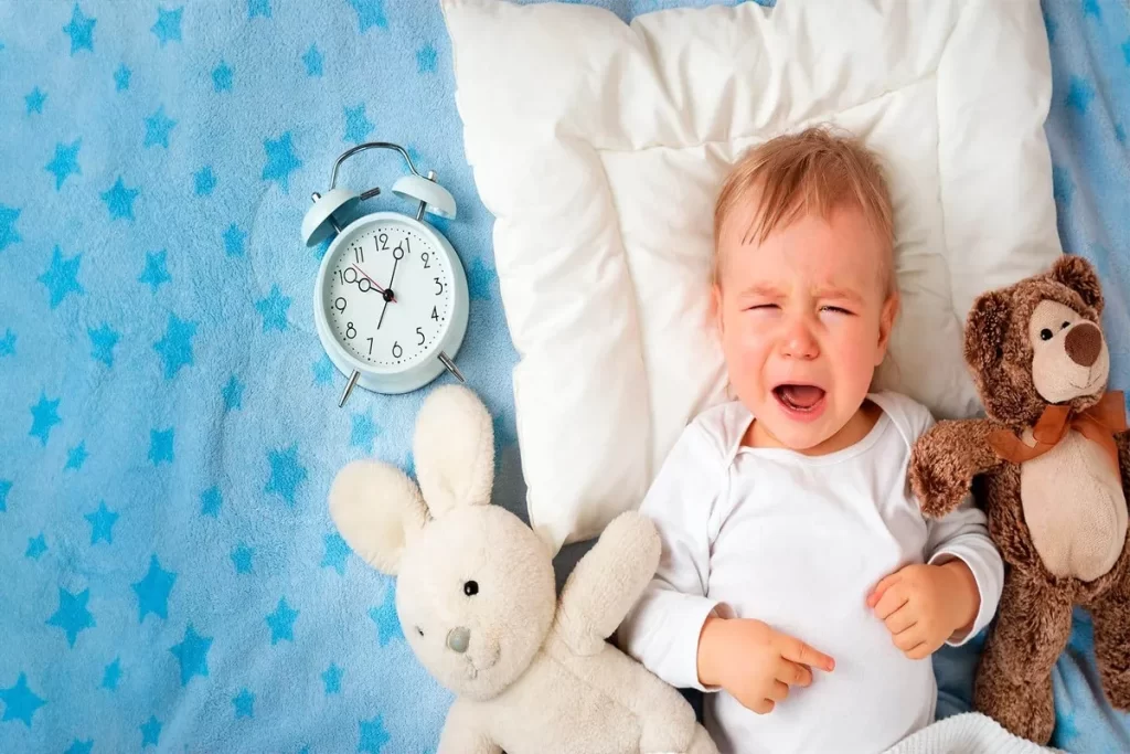 Um bebê chorando deitado de costas na cama, com a boca aberta, em um lençol azul com estrelas brancas. Ao seu lado, estão um despertador e dois bichos de pelúcia: um coelho e um urso. A imagem transmite a aflição do bebê.