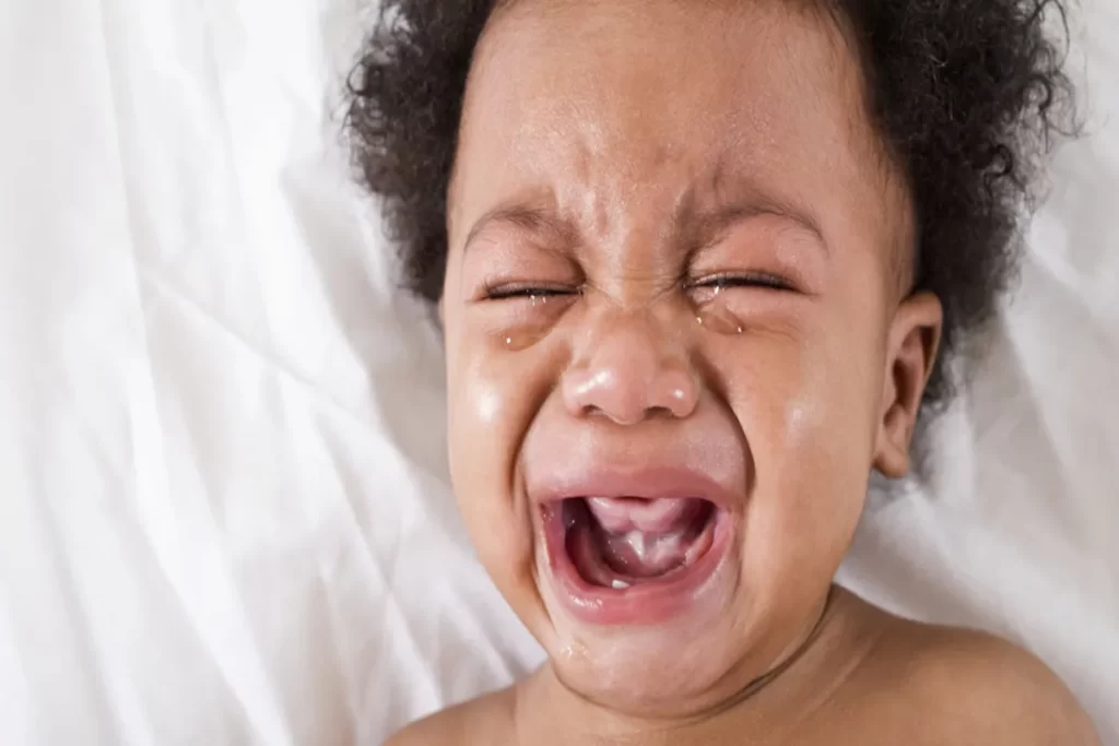 Um bebê com olhos fechados está em uma crise de choro. A boca dele está aberta e há lágrimas escorrendo pelo rosto. A imagem, um close-up, transmite a intensidade e o desespero do choro infantil. O bebê tem cabelo crespo e pele escura. Ele está deitado em um lençol branco, com o foco em seu rosto.