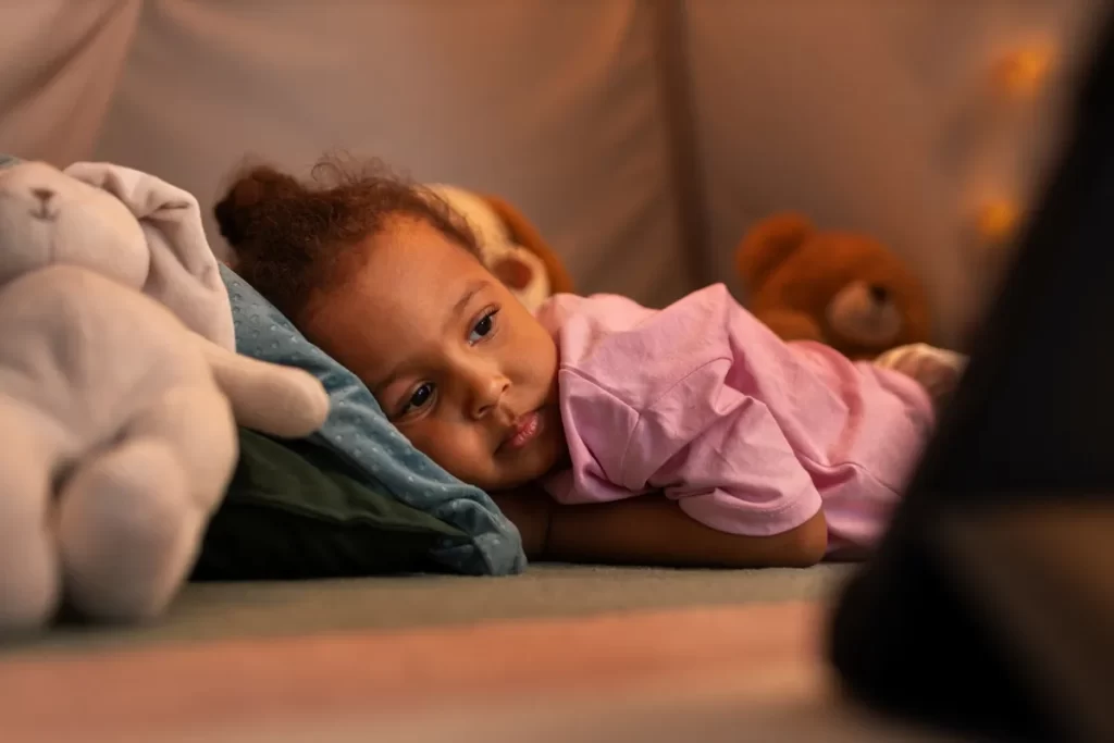 Uma menina pequena deitada em um ambiente aconchegante, com uma expressão triste ou cansada. Ela está cercada por brinquedos de pelúcia, simbolizando a luta contra a insônia e os transtornos do sono na infância.