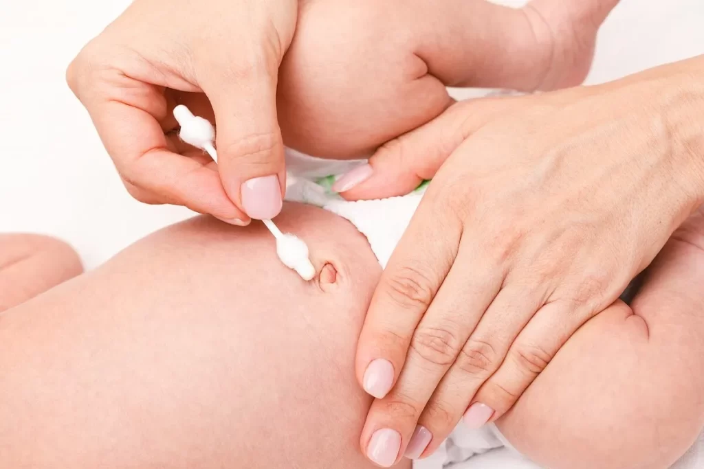 Mãos de um adulto segurando cuidadosamente um cotonete para limpar o umbigo de um bebê deitado em uma superfície branca. A imagem foca nos cuidados preventivos e na higiene do coto umbilical.