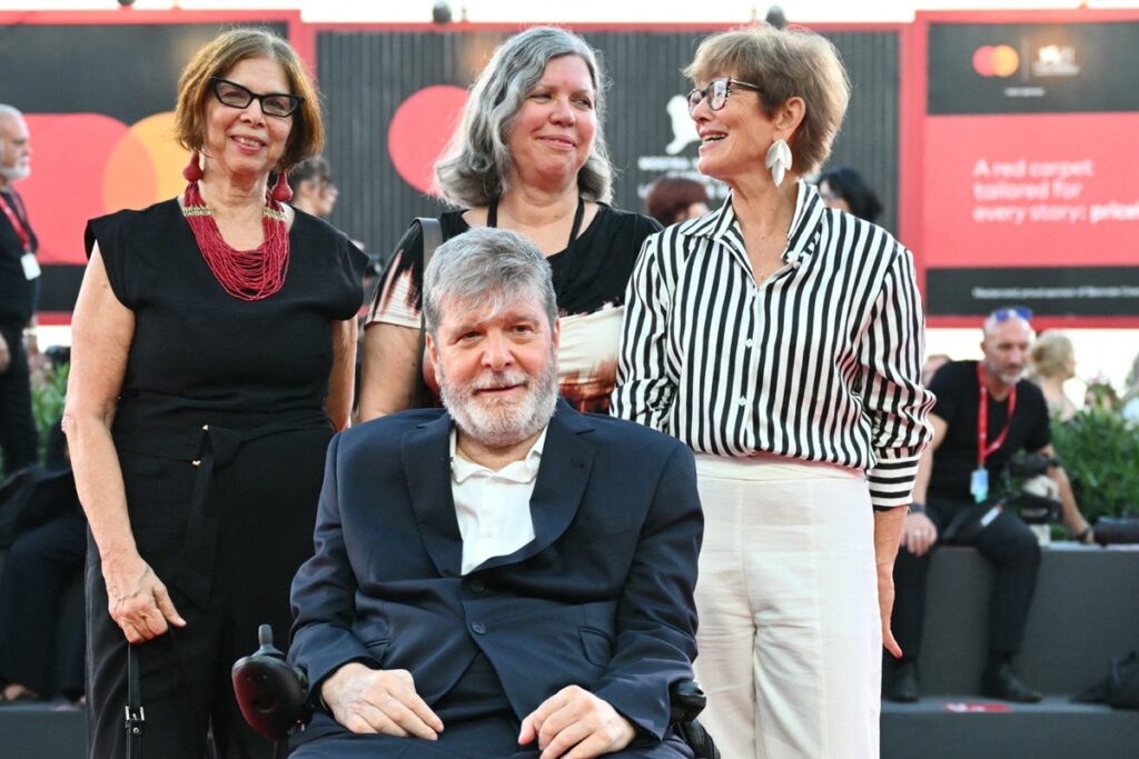Homem de terno azul em cadeira de rodas posando com três mulheres sorridentes em evento público, com pessoas e painéis ao fundo.