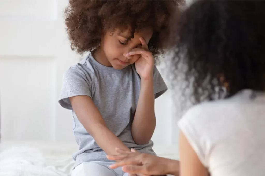 Criança com cabelo cacheado escuro cobrindo o rosto com a mão, sentindo-se angustiada, enquanto uma adulta a conforta.