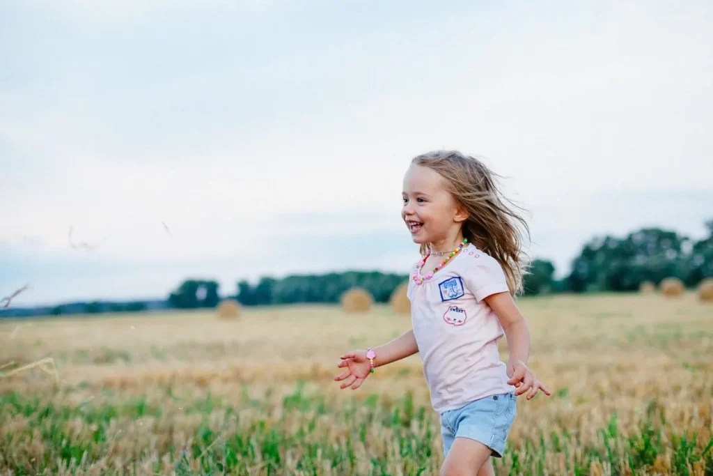 Uma menina de cabelo loiro e comprido, sorridente e cheia de alegria, corre livremente em um campo de feno ou grama seca, com os braços abertos. Ela veste uma camiseta branca e shorts jeans azul-claro. O fundo desfocado (bokeh) mostra grandes rolos de feno (fardos redondos) em um campo rural sob um céu cinza-claro. A imagem simboliza autoconfiança, liberdade e felicidade infantil.