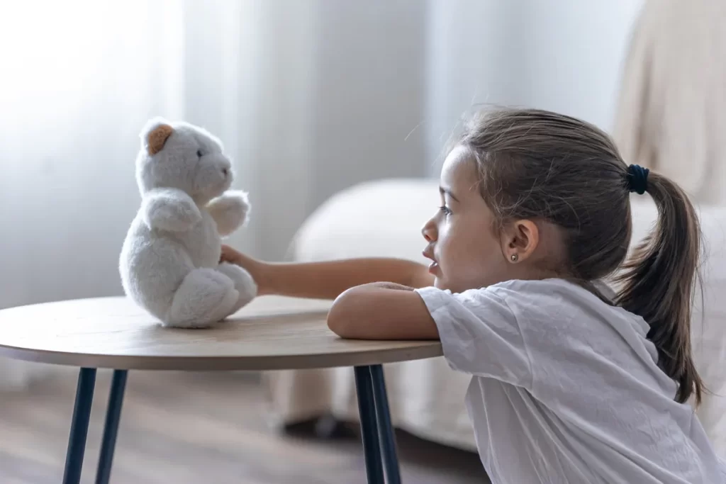 Uma menina de cabelo castanho amarrado em um rabo de cavalo e vestindo uma camiseta branca, está apoiada em uma mesa lateral redonda de madeira clara, interagindo com um urso de pelúcia branco sentado à sua frente. A menina, vista de perfil em um ambiente doméstico e bem iluminado, está concentrada e em uma brincadeira tranquila, simbolizando o foco e o autocontrole.