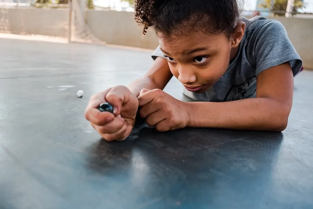 Menina negra com cabelo cacheado deitada no chão, concentrada jogando bolinha de gude ao ar livre.
