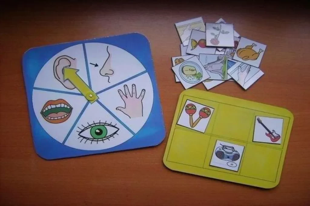 Vista aérea de um jogo de tabuleiro infantil sobre os cinco sentidos, disposto em uma mesa de madeira. O jogo principal é um painel azul redondo com desenhos de uma orelha, nariz, mão, boca e olho, e um ponteiro amarelo. Ao lado, há um painel amarelo com cartas de objetos (maracás, rádio e guitarra) e um monte de cartas de objetos e alimentos (frango, peixe, flor) para associação.