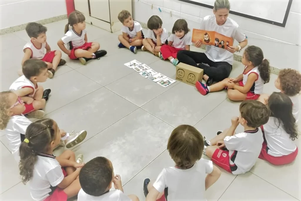 Professora ensinando crianças em idade pré-escolar a identificar emoções com cartões e leitura de um livro em círculo.