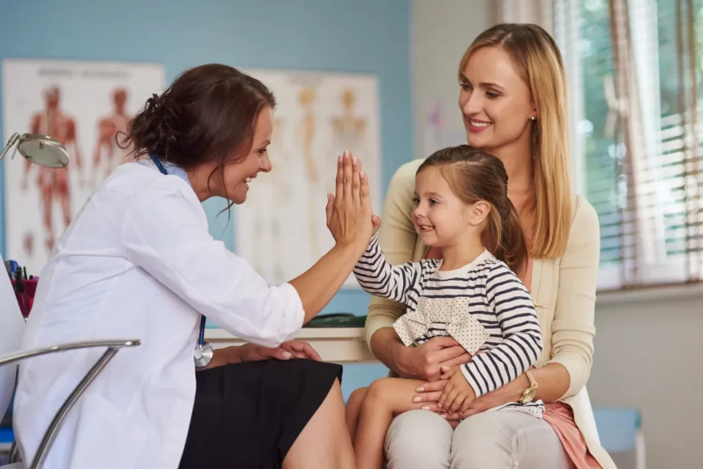 Médica pediatra sorrindo dando um "high five" para uma menina sentada no colo da mãe em um consultório.