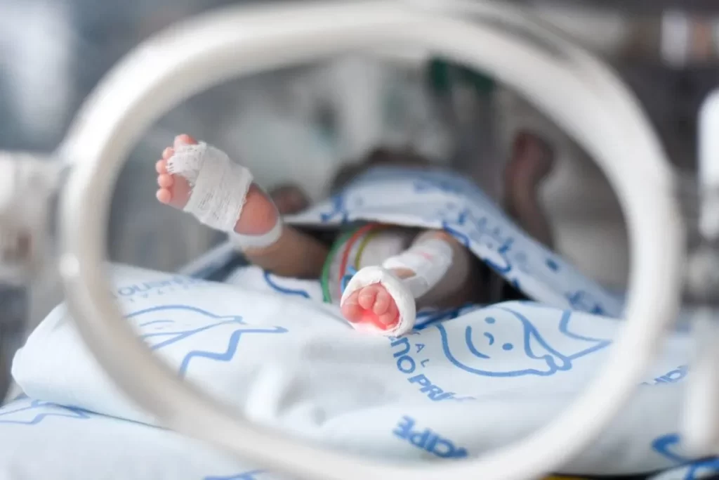 Close-up dos pés de um bebê recém-nascido (com ataduras e sensor de oxímetro) dentro de uma incubadora, vistos através da moldura circular de acesso.