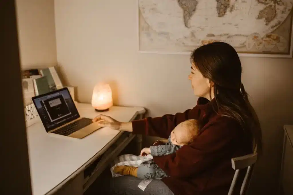 Uma jovem mãe está sentada em uma mesa de trabalho com seu bebê dormindo aninhado em seu colo, enquanto usa um laptop para trabalhar ou estudar em um ambiente doméstico aconchegante.
